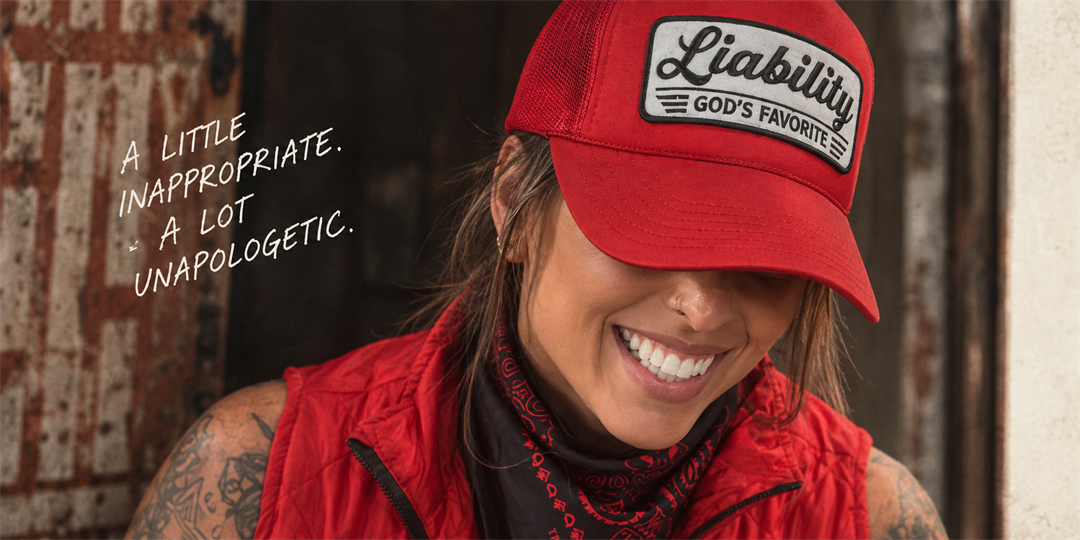 A smiling woman wearing a red Liability God’s Favorite trucker hat